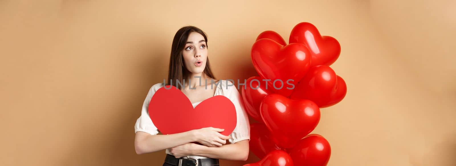 Valentines day and love concept. Intrigued tender girl hugging big red heart cutout and stand near balloons, looking aside at logo, saying wow, beige background.