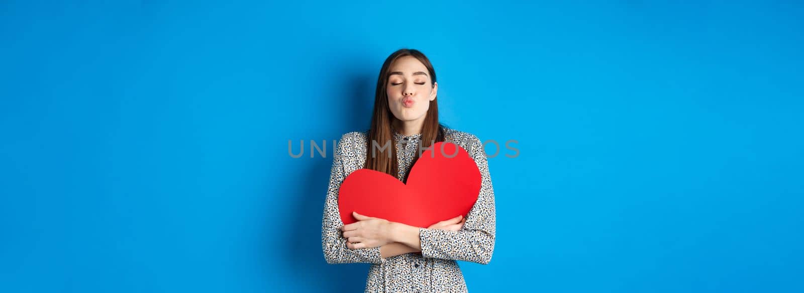 Valentines day. Romantic beautiful woman close eyes and pucker lips for kiss, holding big red heart cutout, kissing you, standing on blue background by Benzoix