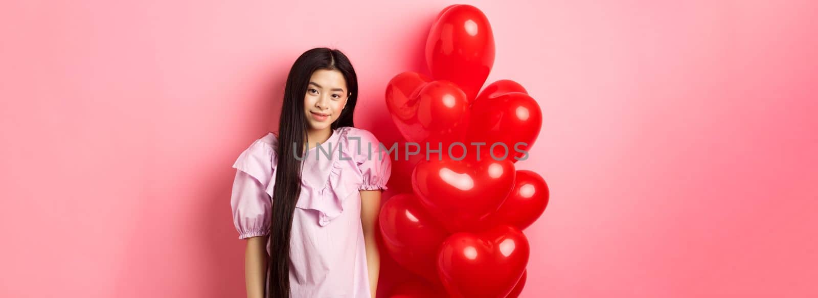 Cute and tender girl looking with love, celebrating valentines and white day with lover, standing near romantic heart balloons on pink background. Relationship concept.