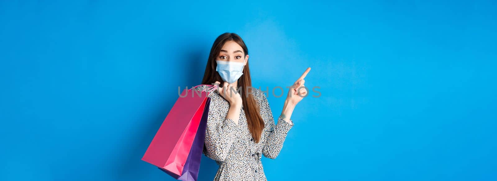 Covid-19, pandemic and lifestyle concept. Excited woman wears medical mask on shopping, pointing left corner logo, holding bags over shoulder, blue background.