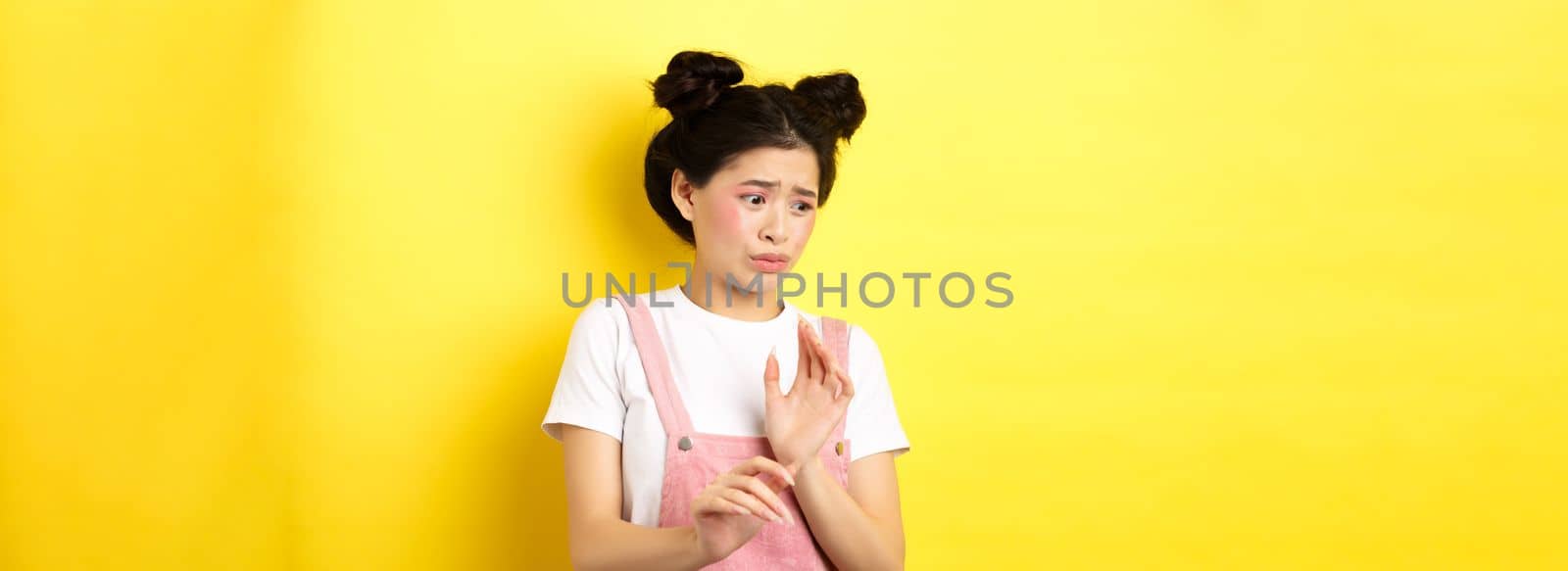 Disgusted asian girl raising hands up defensive, block something disgusting, turn away from aversion and reluctance, standing on yellow background.