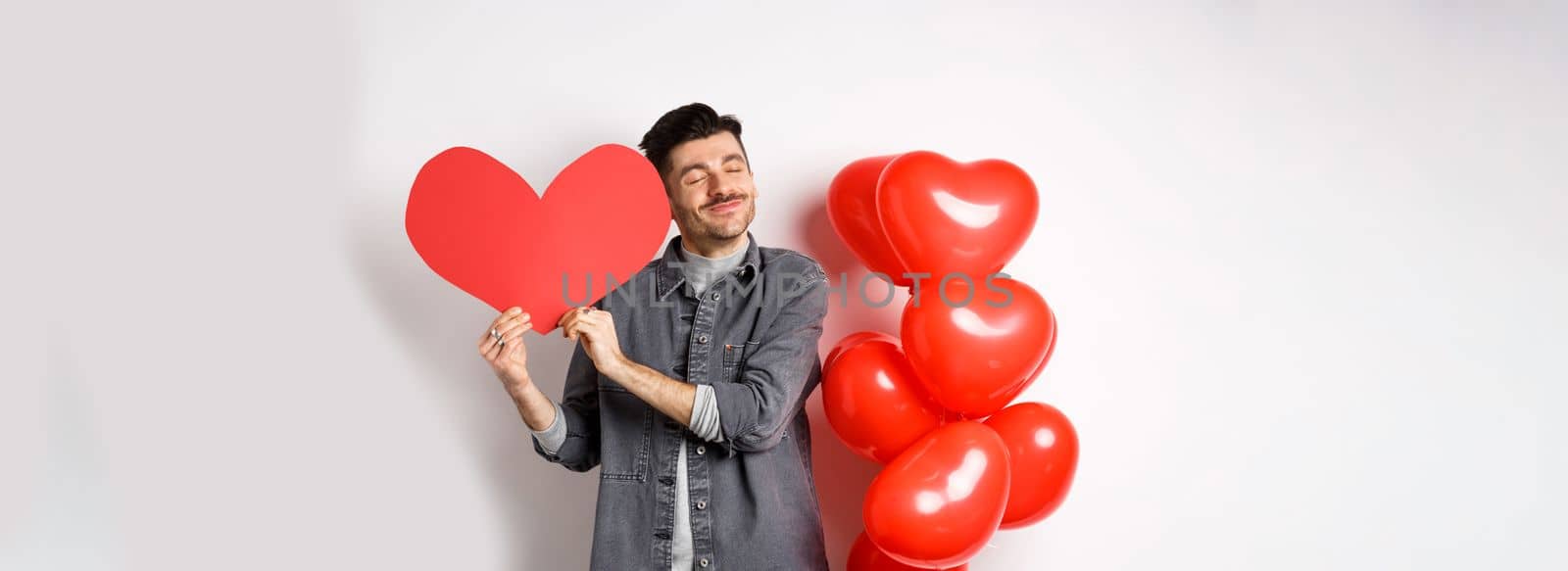 Romantic guy celebrating Valentines day, hugging big red heart card from lover and smiling happy, being in love, standing on white background near red balloons by Benzoix