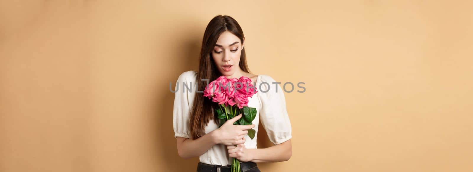 Valentines day. Surprised tender girl looking at beautiful pink roses, receive romantic gift from lover, standing on beige background by Benzoix