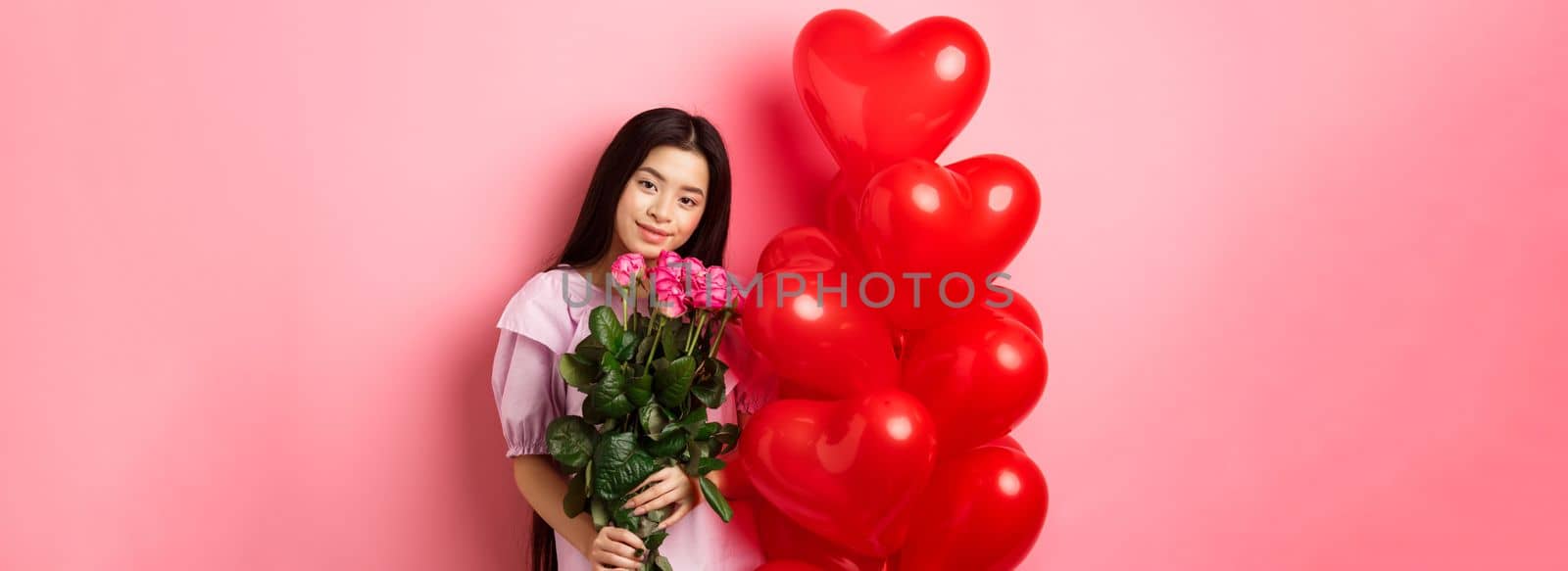 Valentines day concept. Tender and gentle teenage girl holding romantic flowers, standing near red heart balloons from lover and gazing with love, standing on pink background by Benzoix