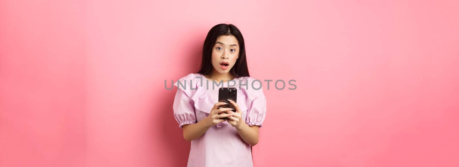 Online shopping. Surprised asian woman in cute dress, open mouth amazed after reading news on smartphone, standing on pink background.