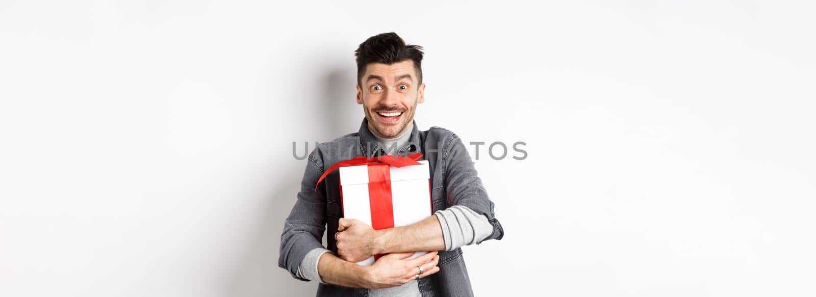 Happy caucasian man holding surprise gift, hugging present and smiling thankful, looking at camera with love and appreciation, celebrate valentines holiday, white background.