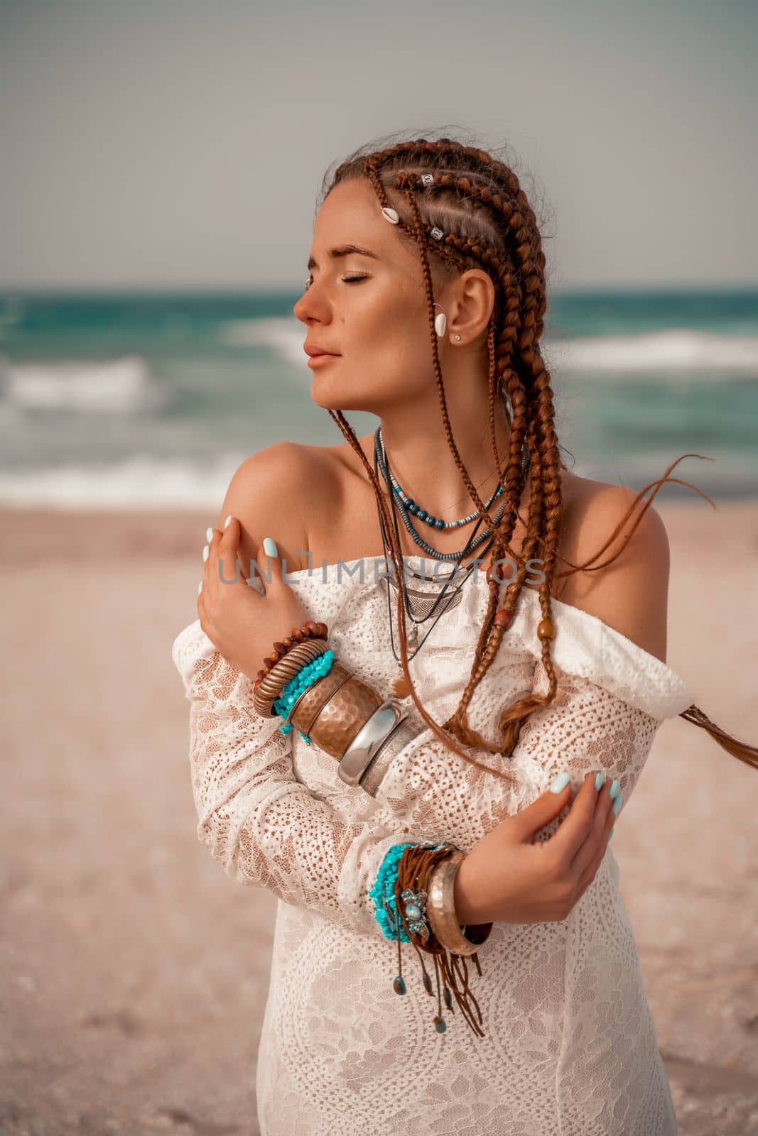 Model in boho style in a white long dress and silver jewelry on the beach. Her hair is braided, and there are many bracelets on her arms