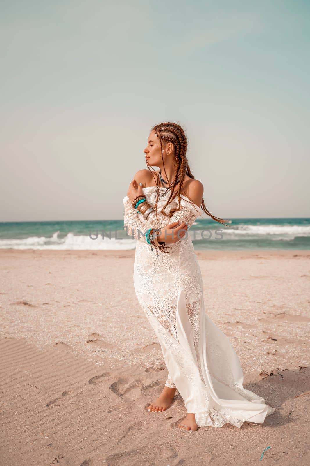 Model in boho style in a white long dress and silver jewelry on the beach. Her hair is braided, and there are many bracelets on her arms