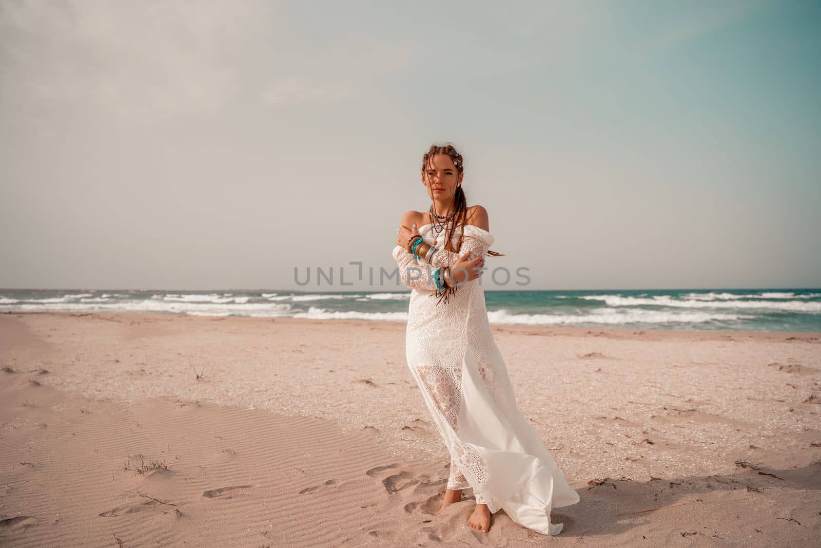 Model in boho style in a white long dress and silver jewelry on the beach. Her hair is braided, and there are many bracelets on her arms