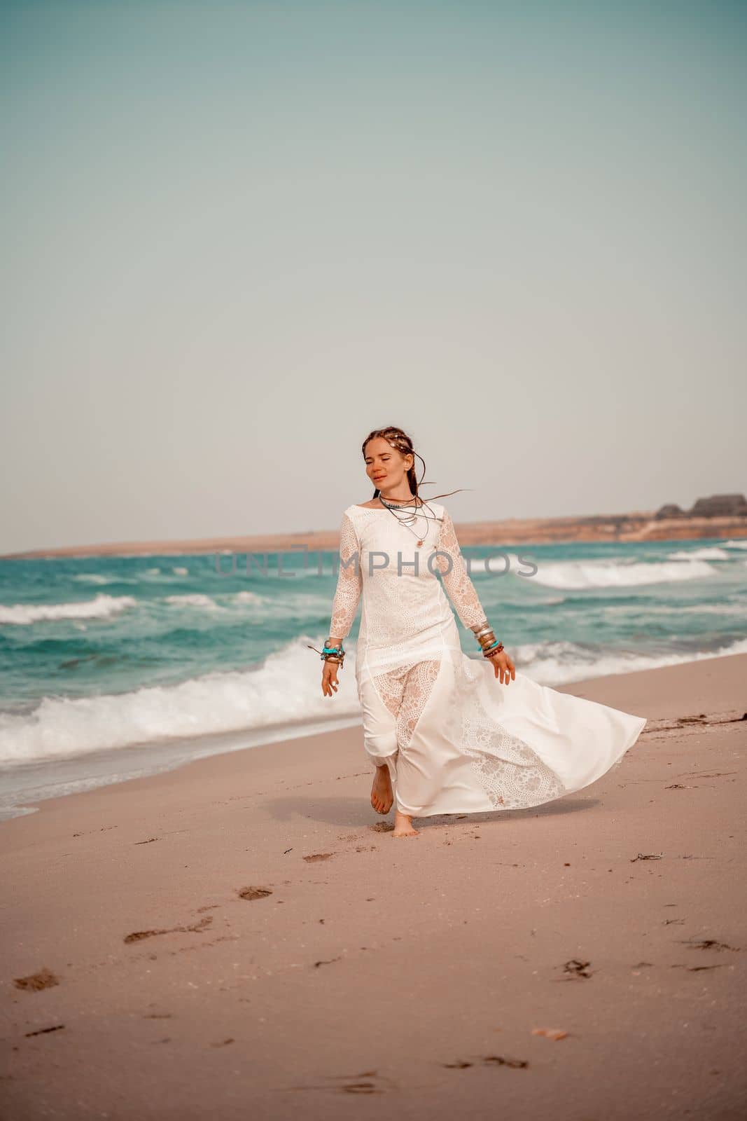 Model in boho style in a white long dress and silver jewelry on the beach. Her hair is braided, and there are many bracelets on her arms