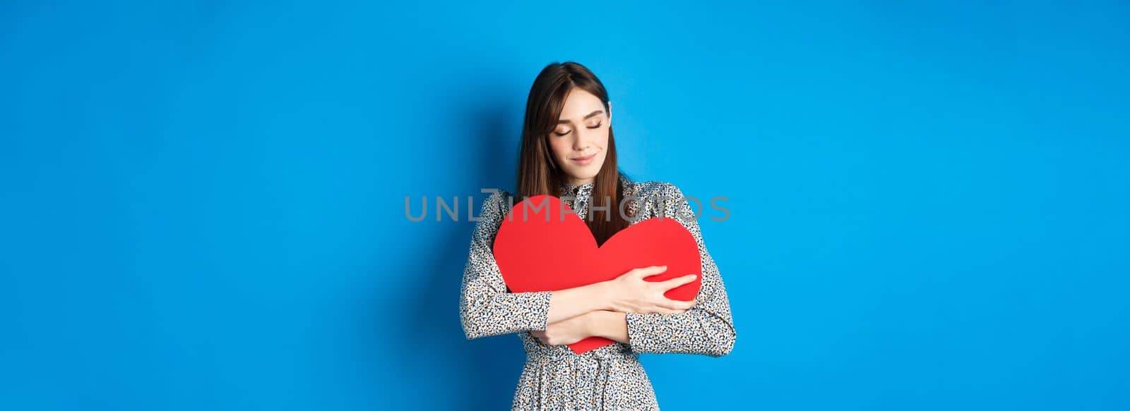 Valentines day. Romantinc girl in dress hugging big red heart cutout, close eyes and smile with dreamy face, imaging sensual date, standing on blue background.