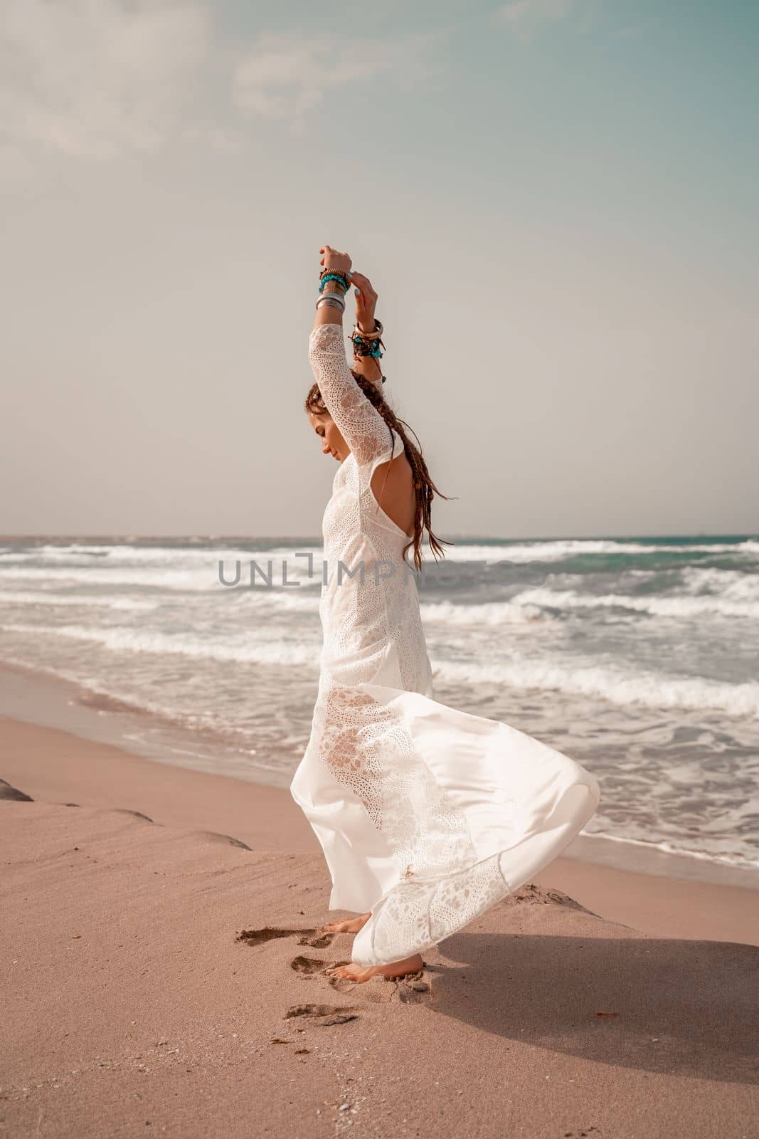 Model in boho style in a white long dress and silver jewelry on the beach. Her hair is braided, and there are many bracelets on her arms