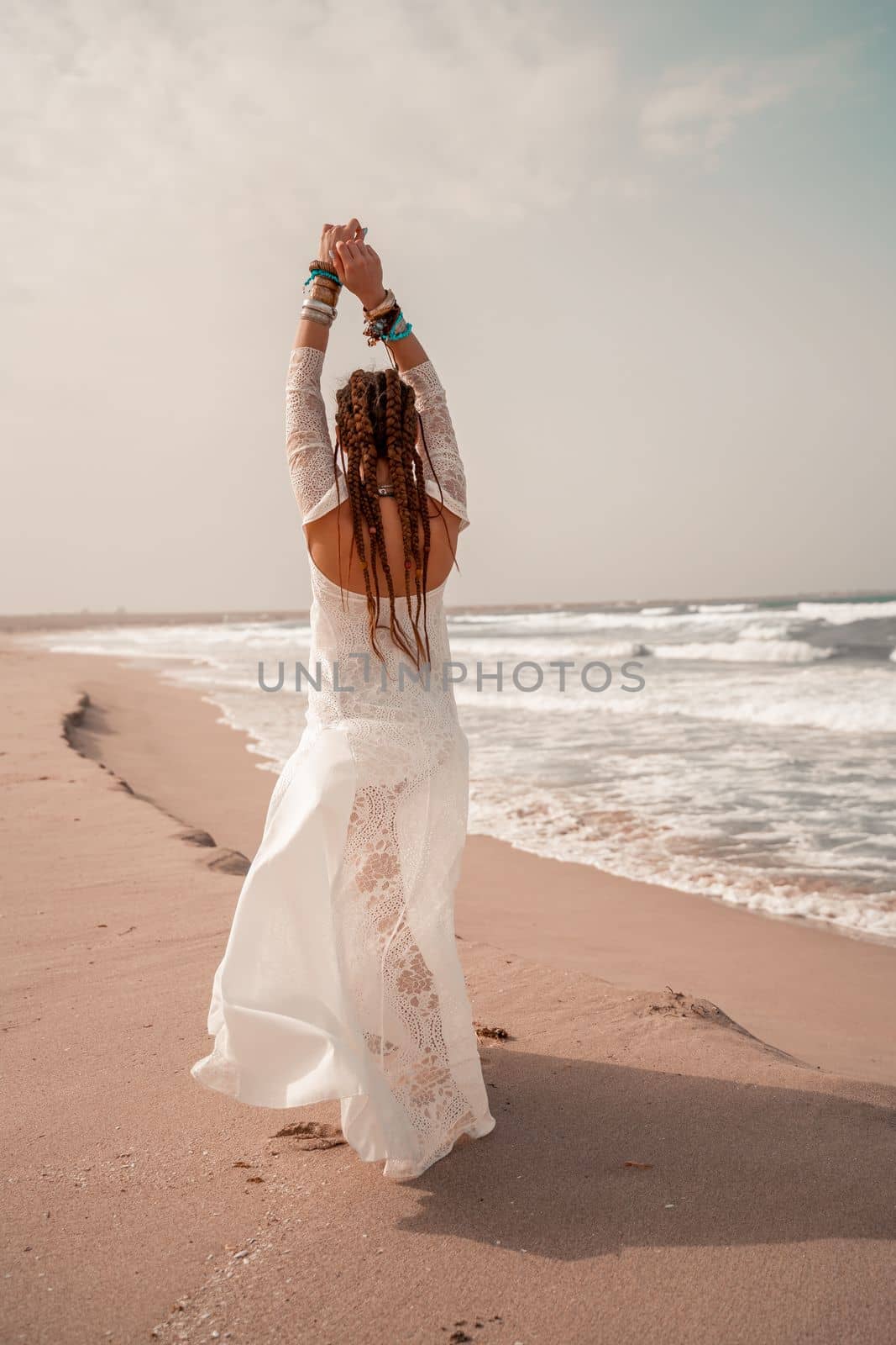 Model in boho style in a white long dress and silver jewelry on the beach. Her hair is braided, and there are many bracelets on her arms