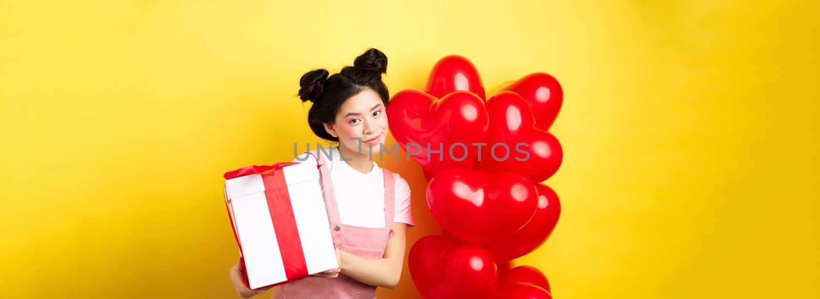 Happy Valentines day. Beautiful and stylish asian woman posing near heart balloons with lovely box with gift from lover, standing over yellow background.