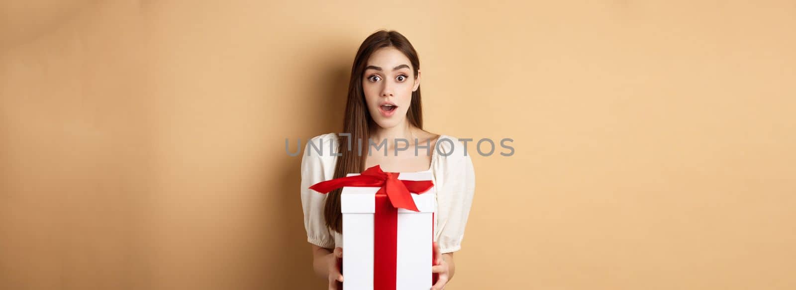 Amazed beautiful girl receiving gift from secret admirer on Valentines day, looking surprised at camera, holding box and standing on beige background by Benzoix