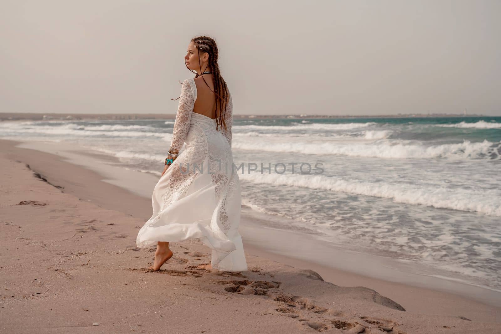 Model in boho style in a white long dress and silver jewelry on the beach. Her hair is braided, and there are many bracelets on her arms