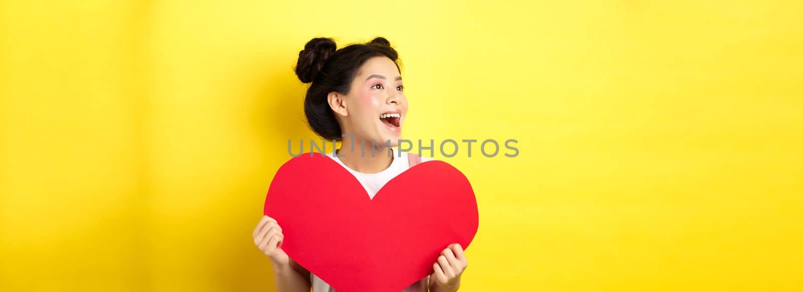 Happy valentines and love concept. Cheerful japanese girl looking left, showing big red heart and smiling happy at lover, standing on yellow background.