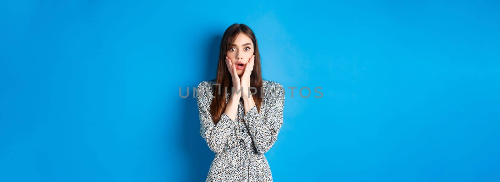 Shocked cute woman gasping amazed, drop jaw and stare with disbelief, standing in dress on blue background.