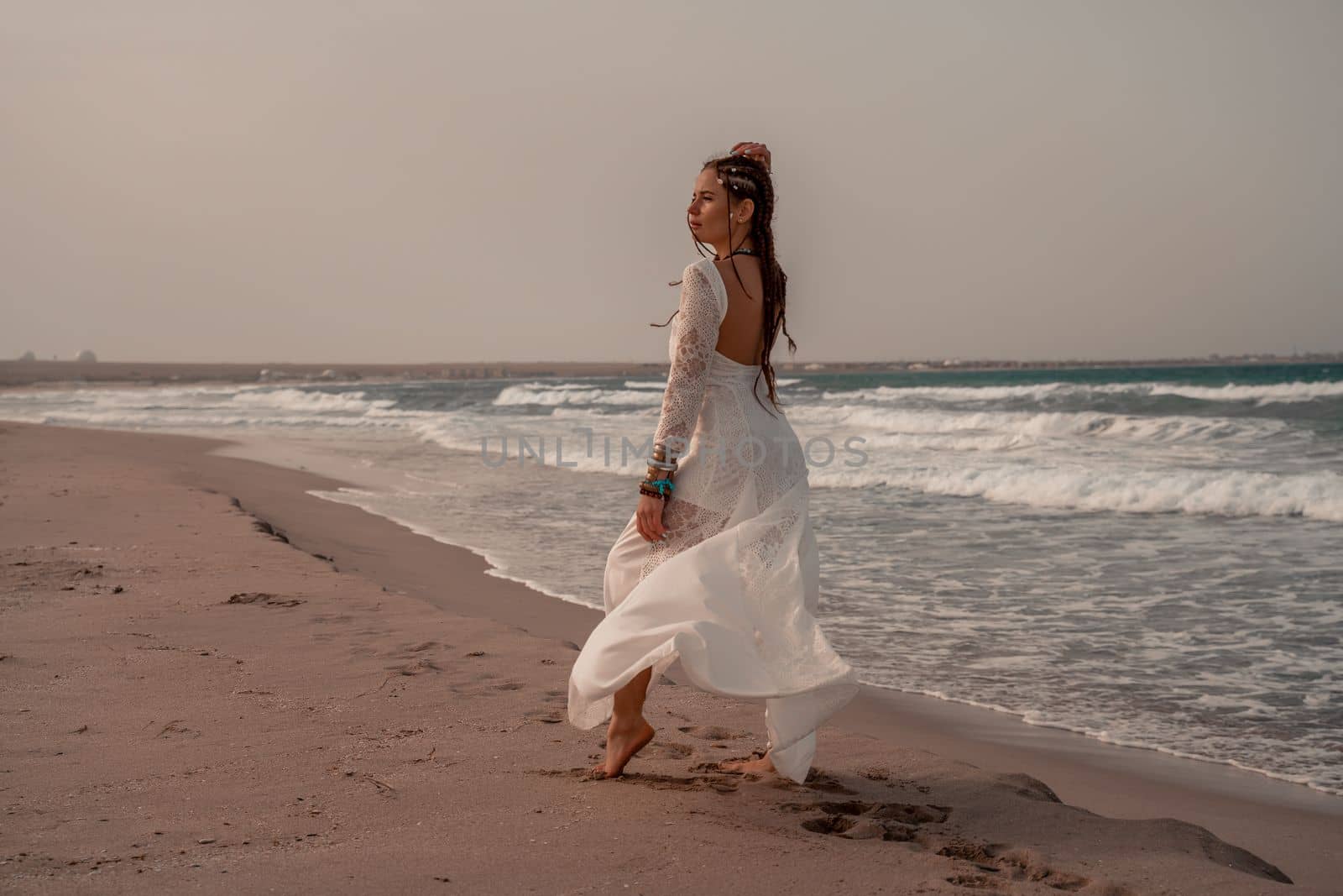 Model in boho style in a white long dress and silver jewelry on the beach. Her hair is braided, and there are many bracelets on her arms