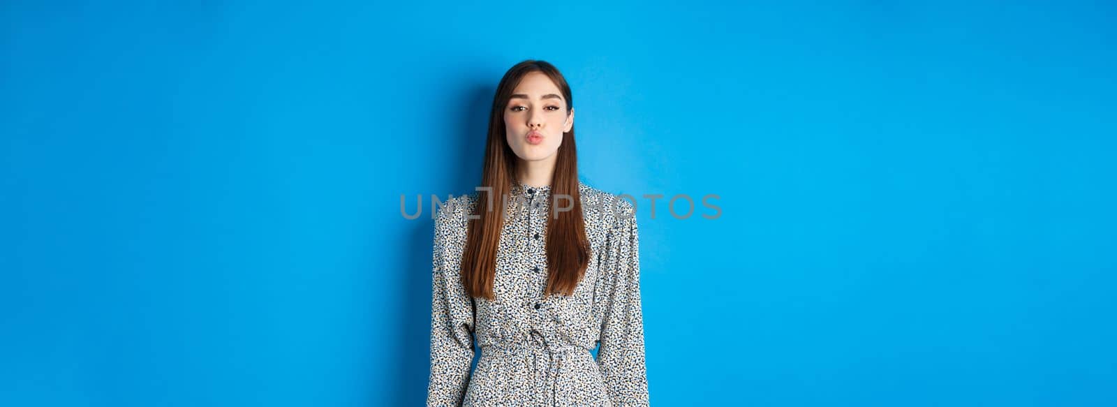 Valentines day and relationship concept. Lovely woman in dress pucker lips for kiss, looking with love at at camera, standing on blue background.