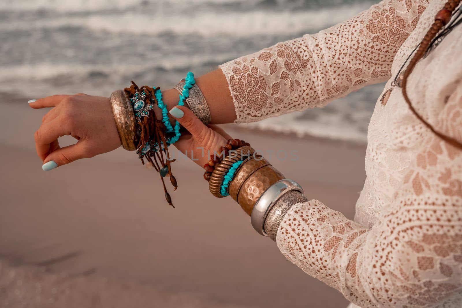 Model in boho style in a white long dress and silver jewelry on the beach. Her hair is braided, and there are many bracelets on her arms