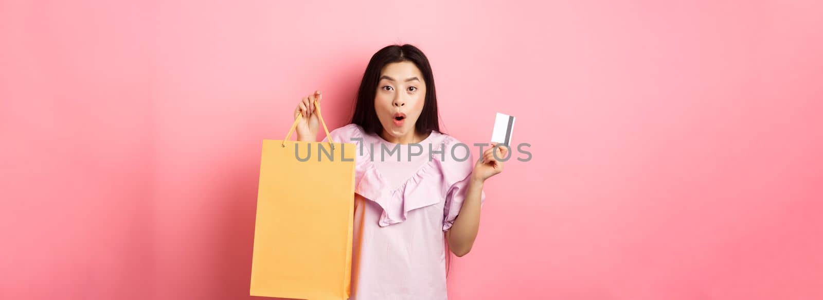 Shopping. Excited asian woman gasping amazed, holding bag from shop and plastic credit card, standing on pink background.