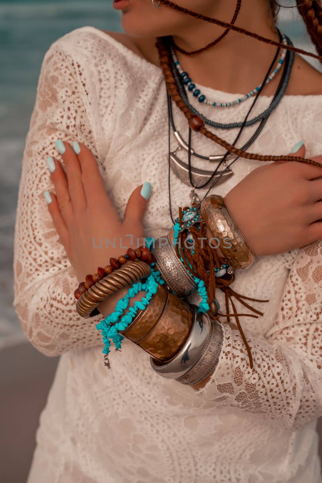 Model in boho style in a white long dress and silver jewelry on the beach. Her hair is braided, and there are many bracelets on her arms