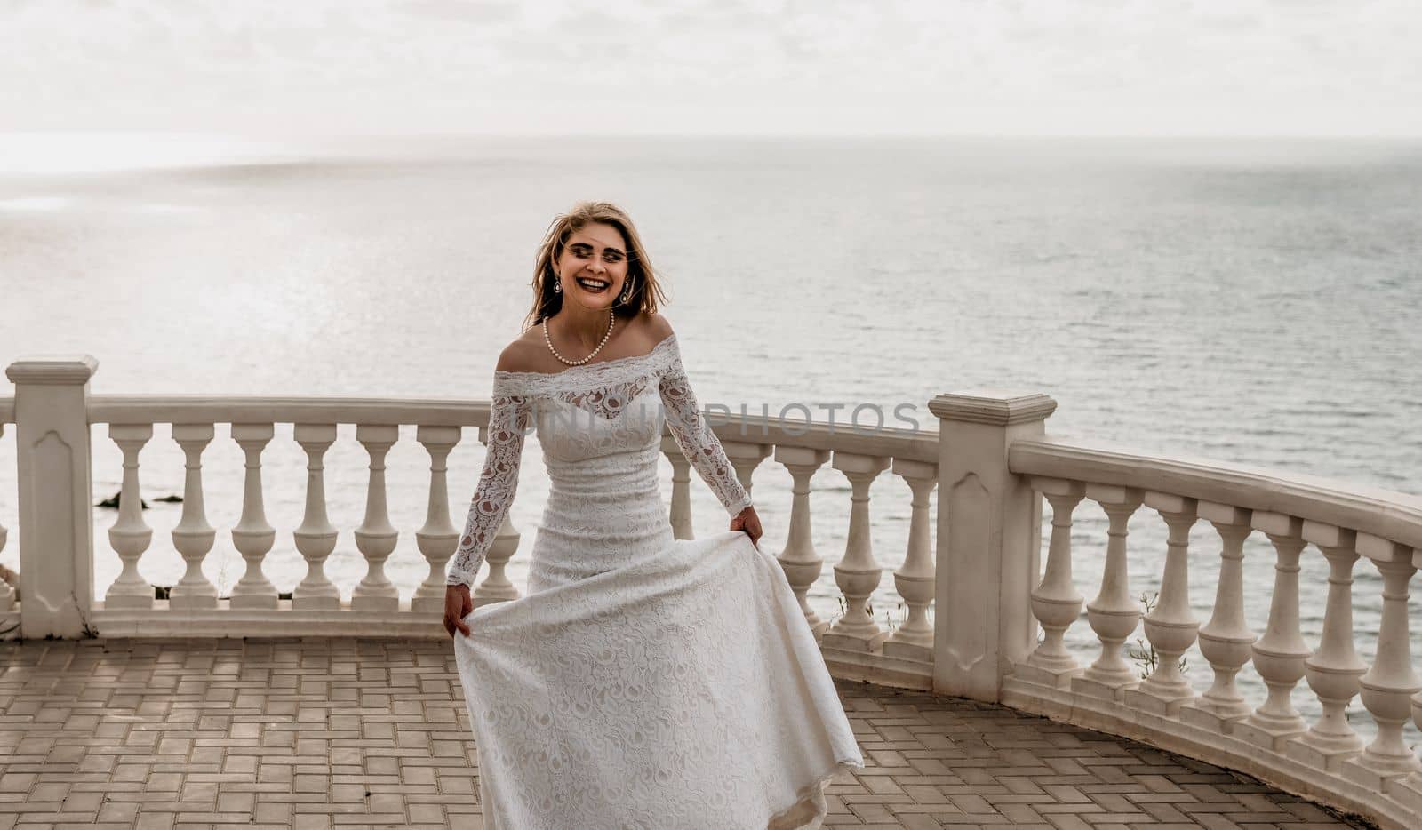 Romantic bride, blond girl in white wedding dress with open shoulders posing on open terrace with backdrop of the sea and rocks. Stylish young woman standing on terrace and looking on ocean at sunset by panophotograph