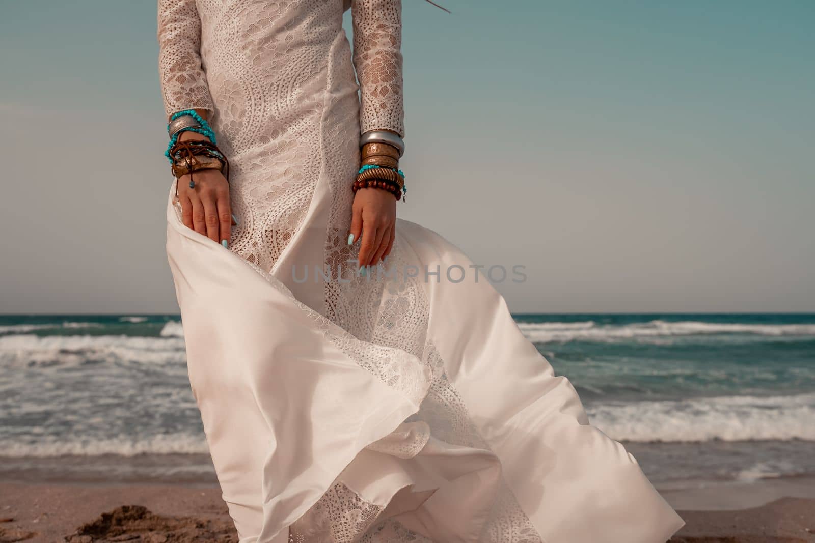 Model in boho style in a white long dress and silver jewelry on the beach. Her hair is braided, and there are many bracelets on her arms