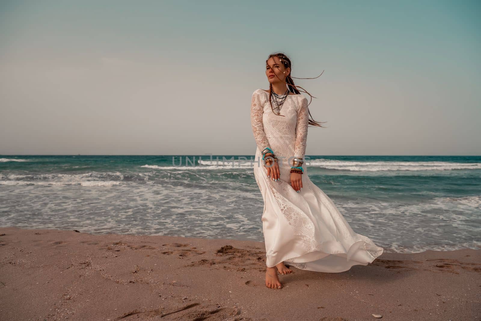 Model in boho style in a white long dress and silver jewelry on the beach. Her hair is braided, and there are many bracelets on her arms