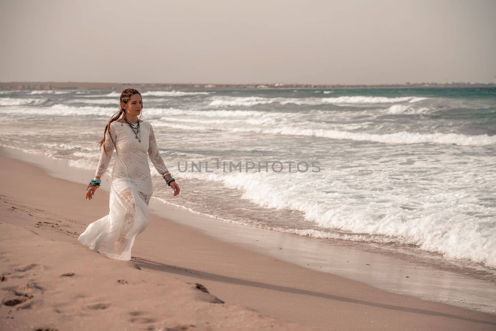 Model in boho style in a white long dress and silver jewelry on the beach. Her hair is braided, and there are many bracelets on her arms