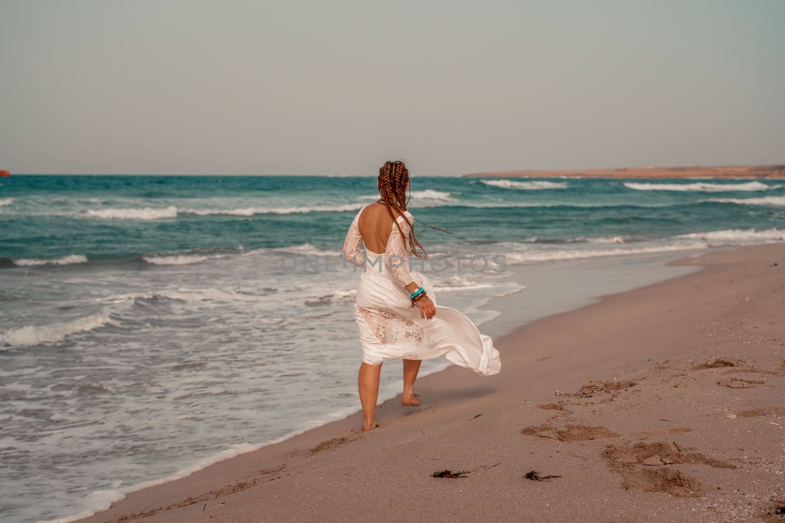 Model in boho style in a white long dress and silver jewelry on the beach. Her hair is braided, and there are many bracelets on her arms