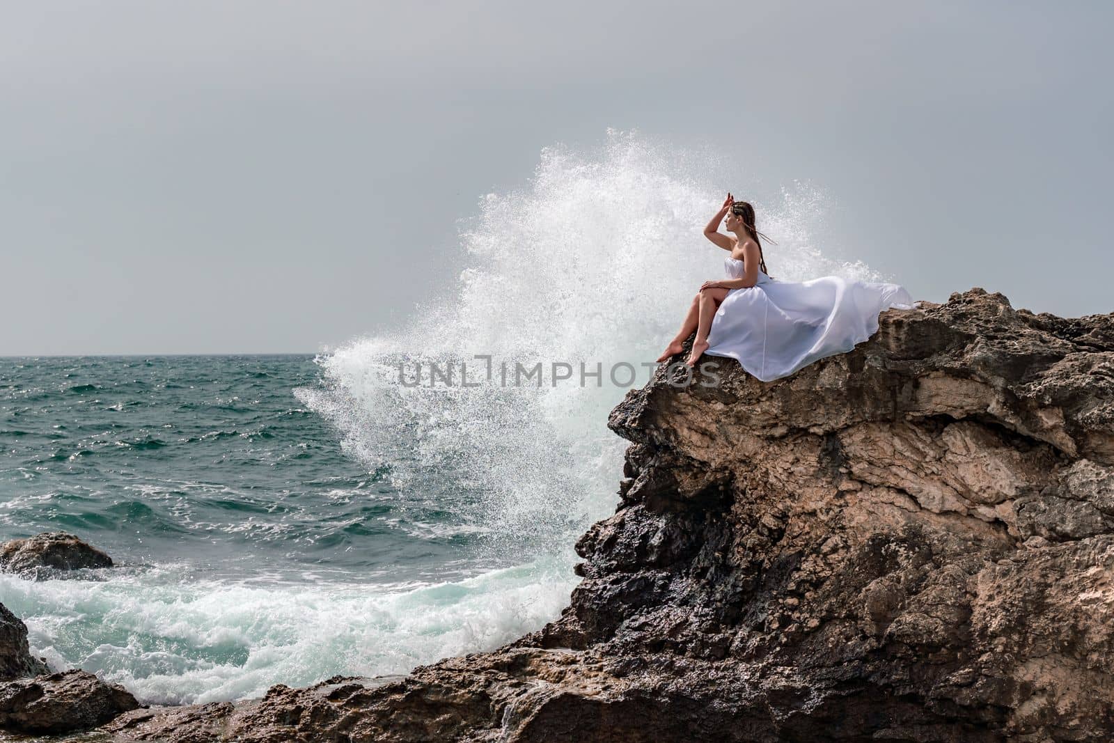 A woman in a storm sits on a stone in the sea. Dressed in a white long dress, waves crash against the rocks and white spray rises above her