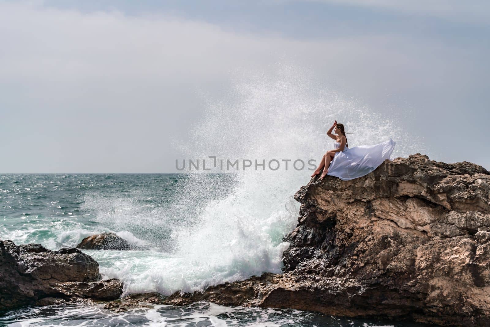 A woman in a storm sits on a stone in the sea. Dressed in a white long dress, waves crash against the rocks and white spray rises above her