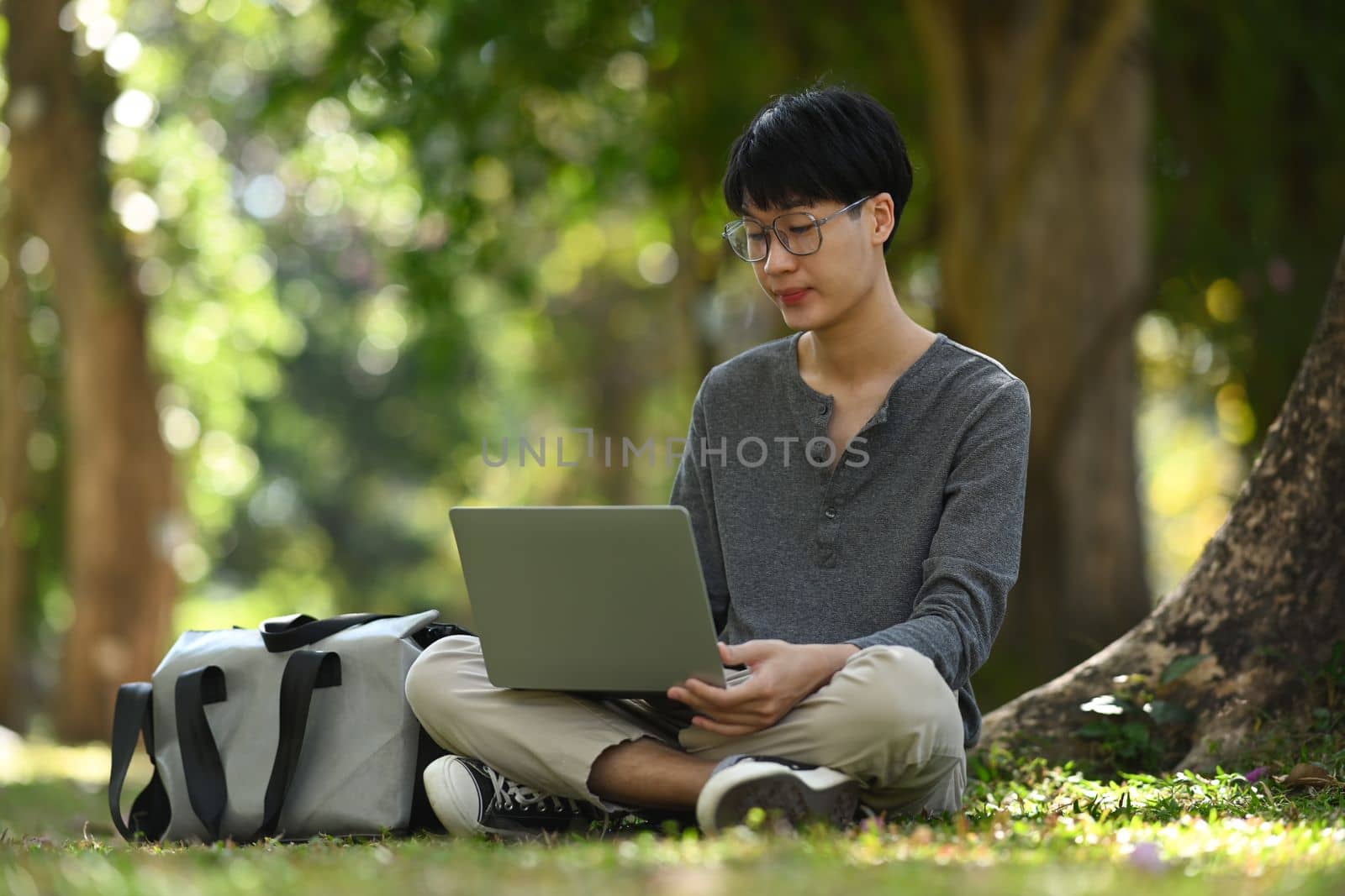 Casual asian man student using laptop while sitting on green grass in city park. Education, technology and lifestyle concept.