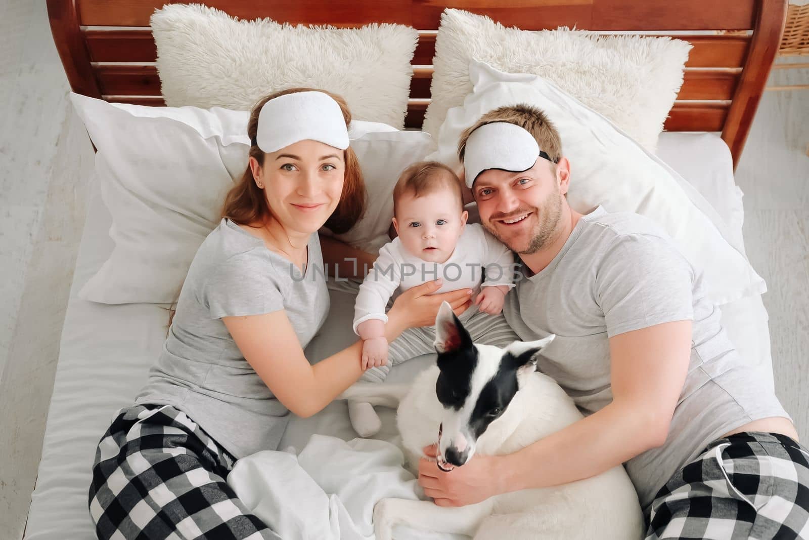 Family with child and dog lying in the bed together, looking at the camera and smiling. Young mother and father resting with toddler kid and pet doggy in the morning