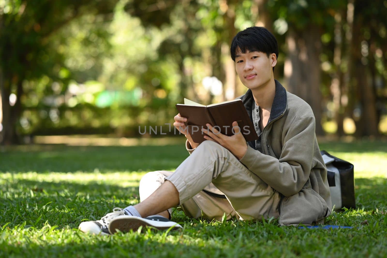 Pleasant asian man reading favorite book on a green meadow in beautiful summer day.