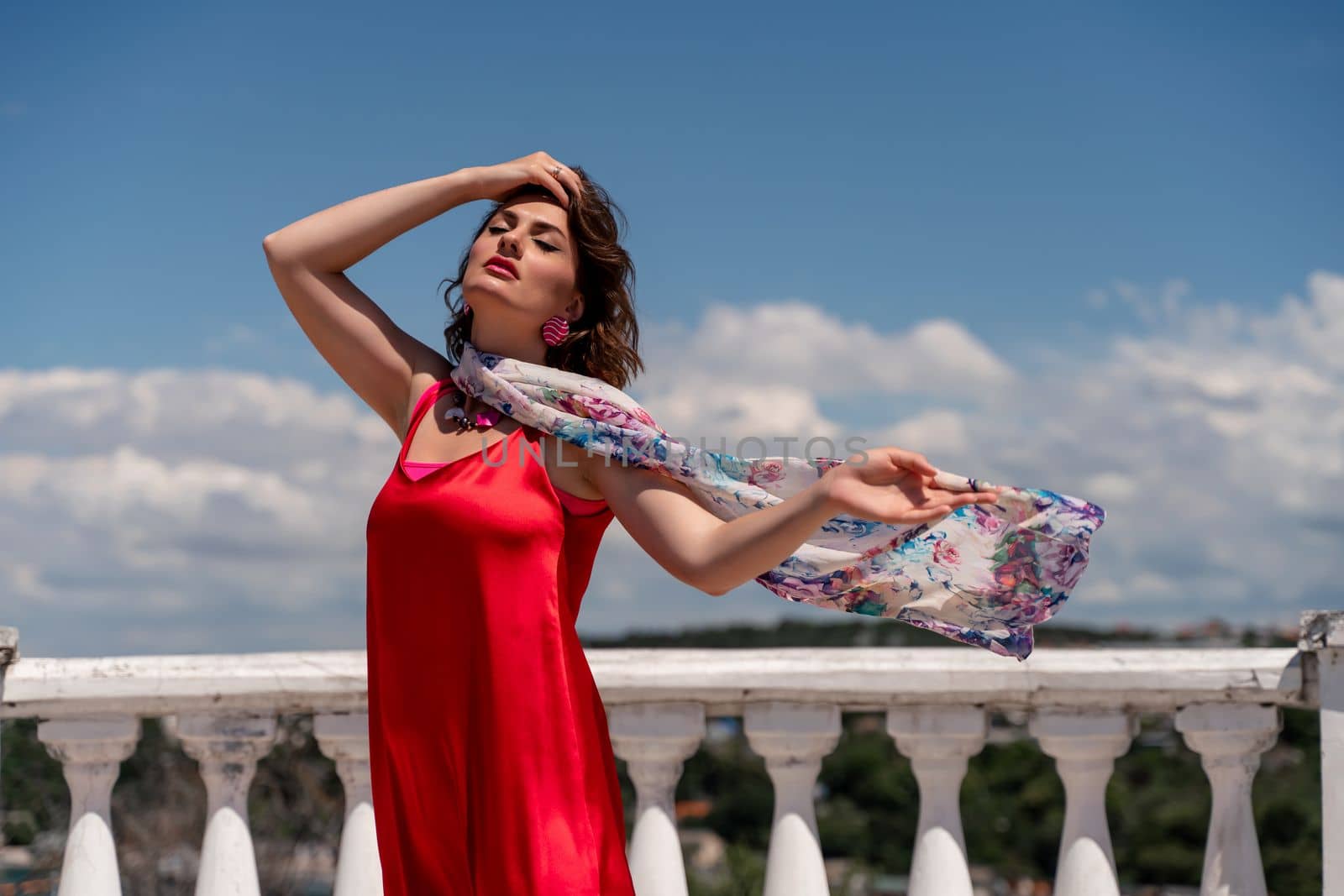 A woman in a red dress against a blue sky and white clouds with a developing scarf around her neck. She turned her face to the wind and posed for the camera. by Matiunina