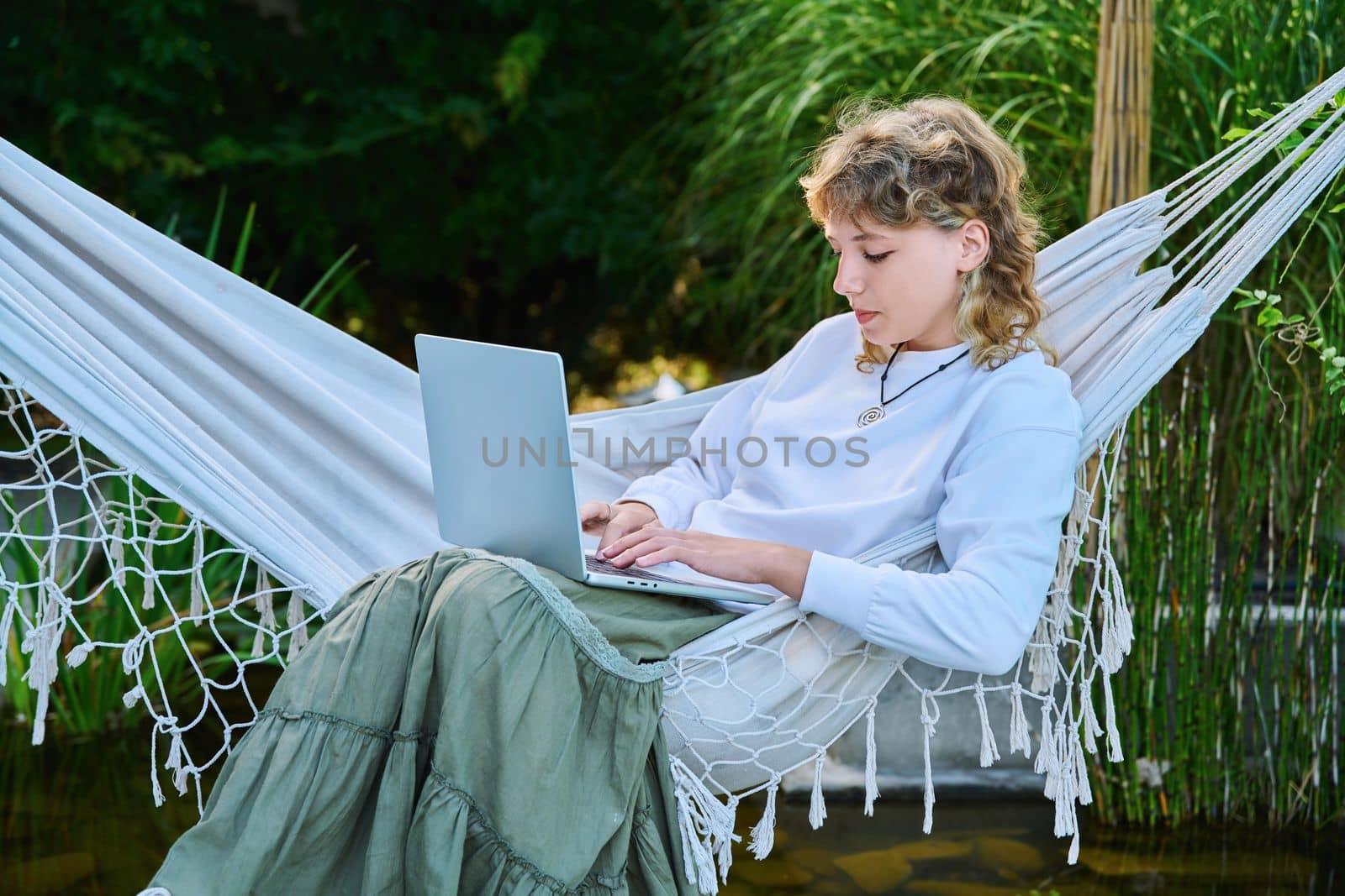 Teenage girl relaxing in hammock using laptop for leisure study by VH-studio