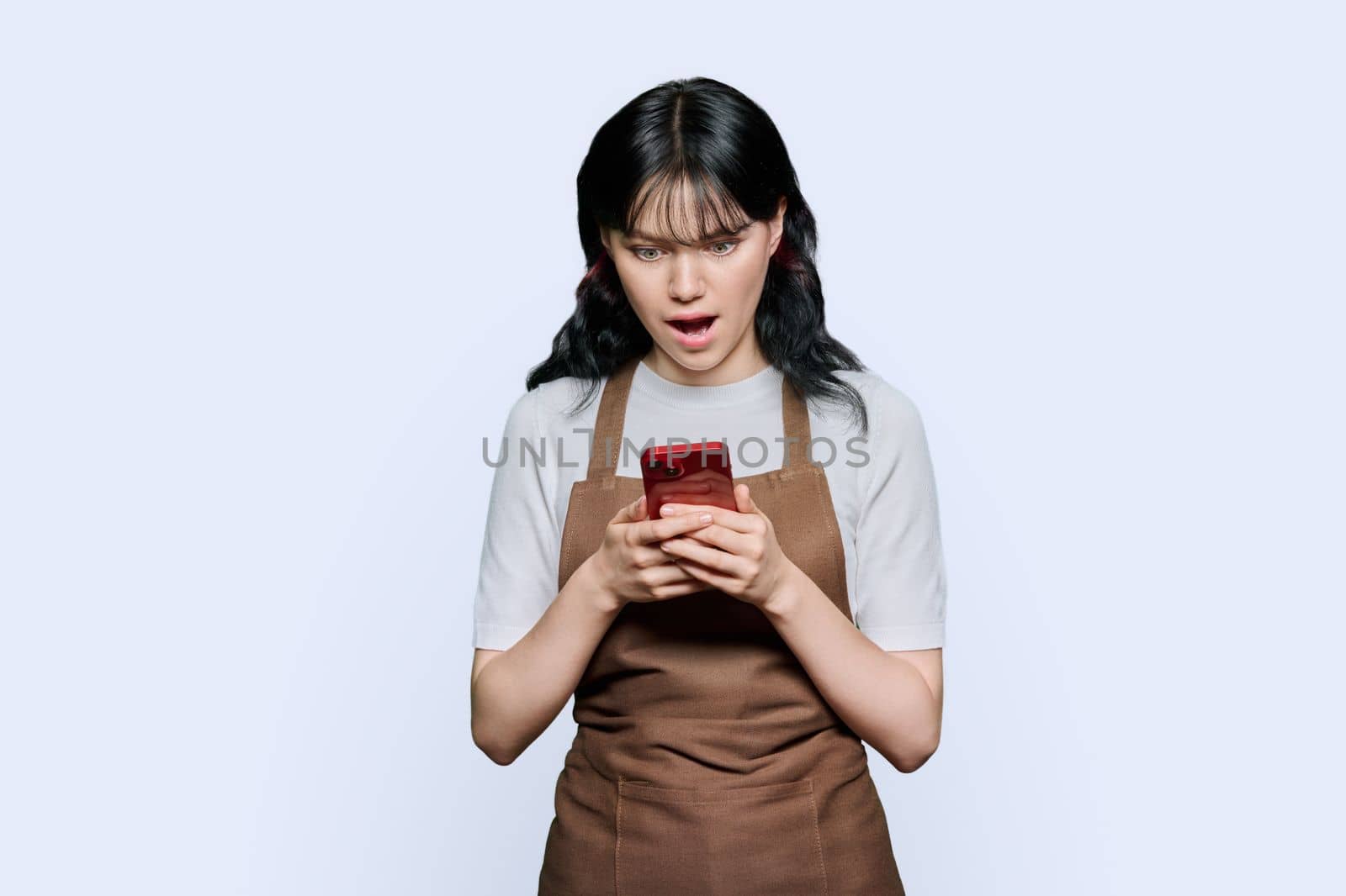 Shocked emotional young woman in apron looking surprised at smartphone screen on white studio background