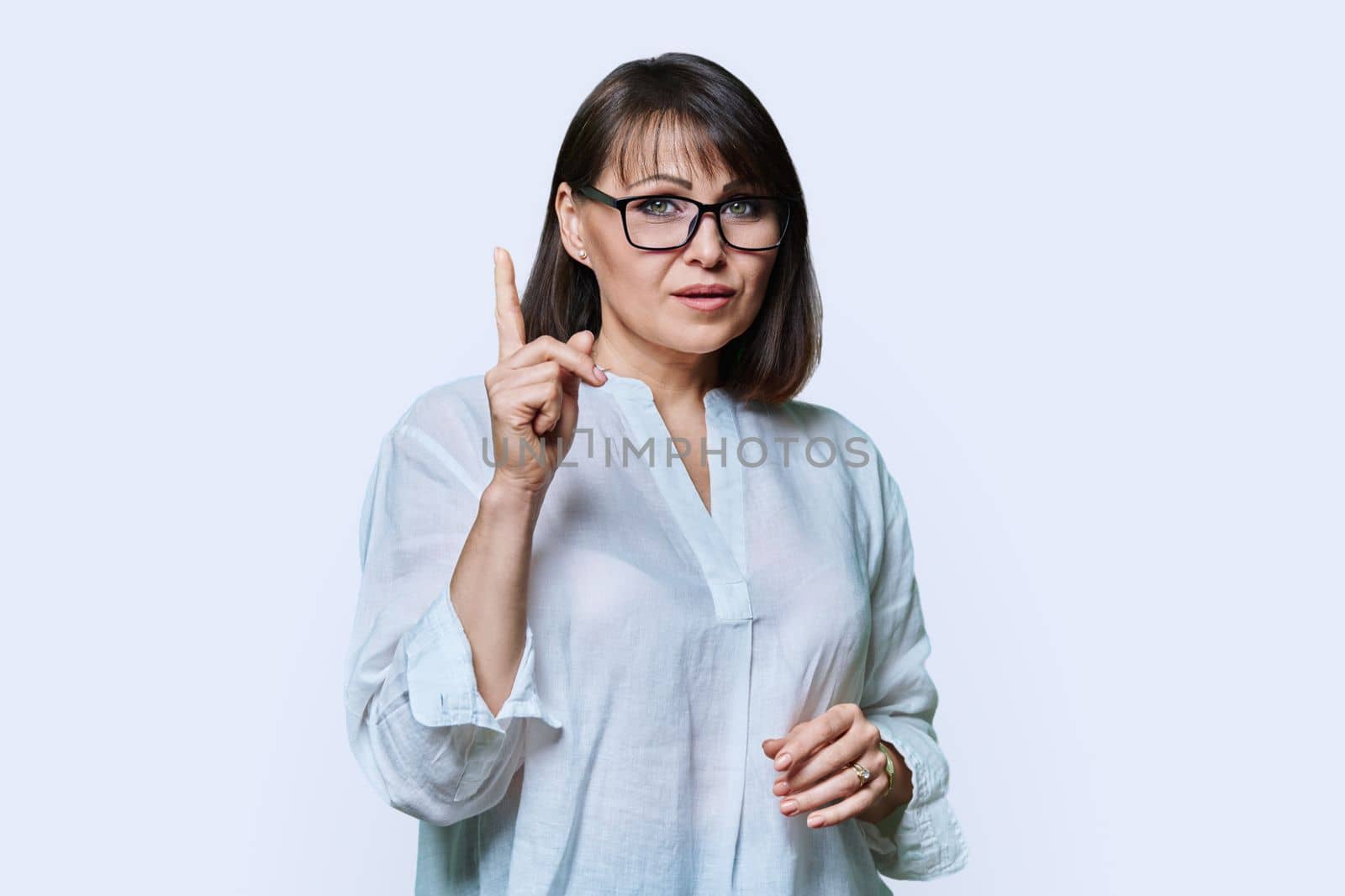 Middle aged serious woman showing index finger up, looking at camera, attention sign, on white studio background