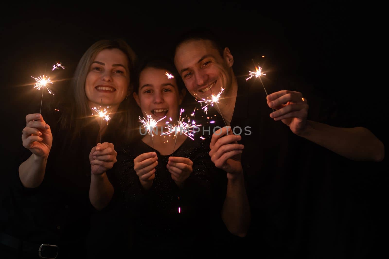 Happy family in black - woman, man and little girl, with a sparkler in hand the New Year's Christmas at home. The girl at the father on hands. New year 2023. by Andelov13