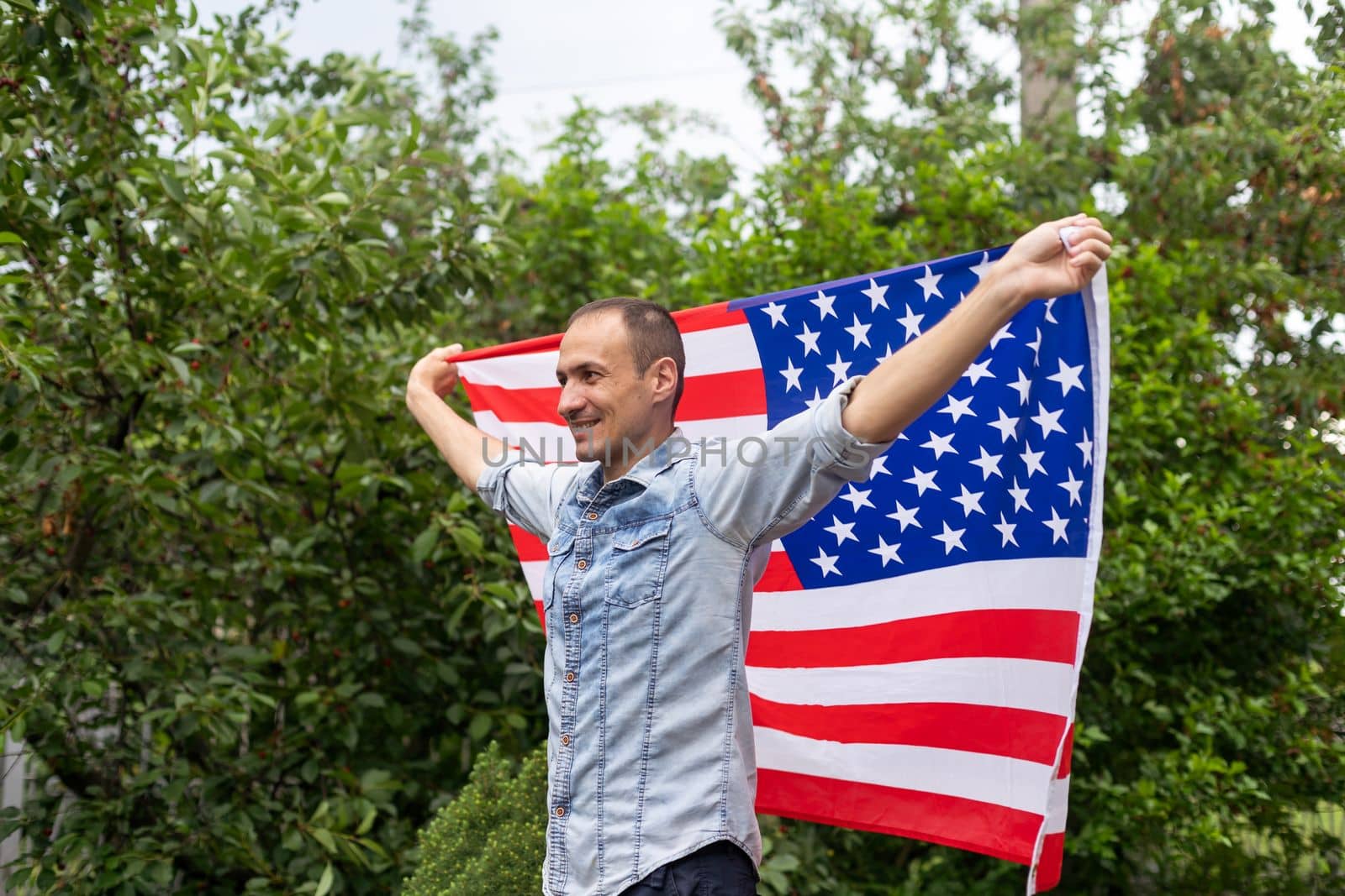 Positive attractive american young guy wrapped himself in the flag of America and looks at the camera, outdoors. High quality photo