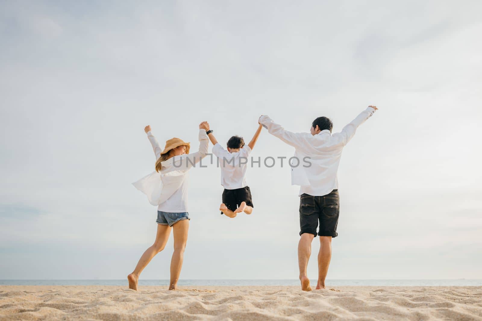 Happy Asian family have fun and live healthy lifestyle together on beach, Back family mother, father and son holding hands and jumping in air at dawn time, Family outdoor activities concept