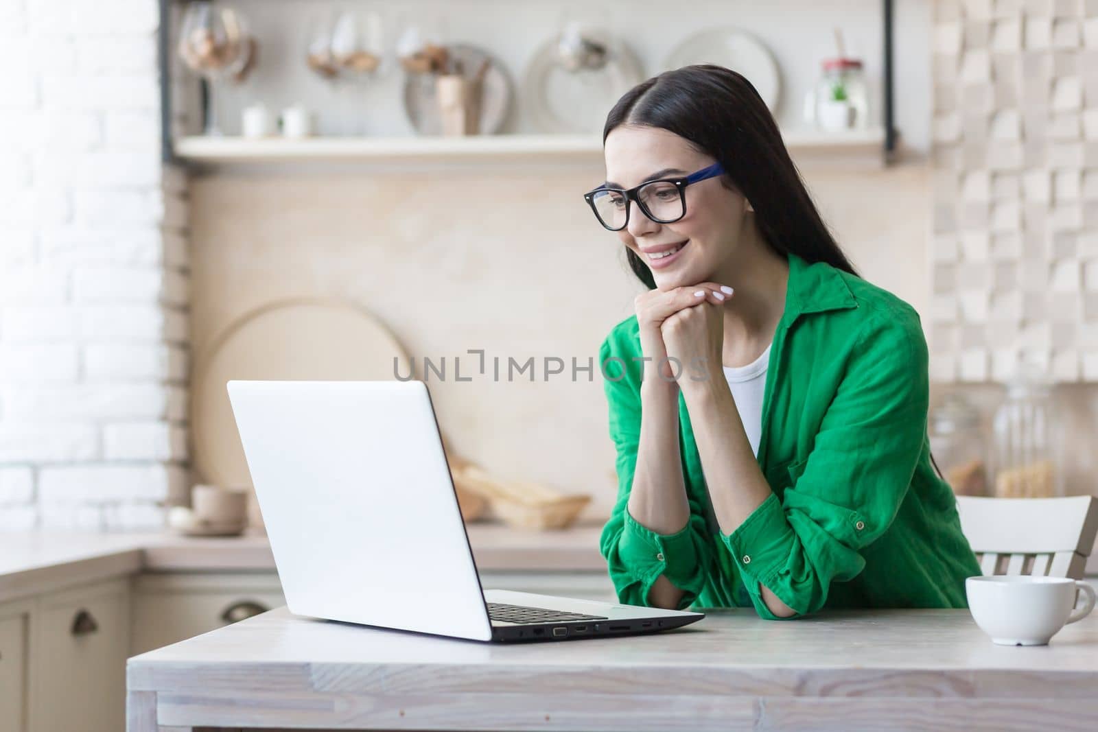 Young beautiful female blogger sitting at home in kitchen and chatting online on laptop camera. She rested her head on her hands, listens, tells stories, smiles.