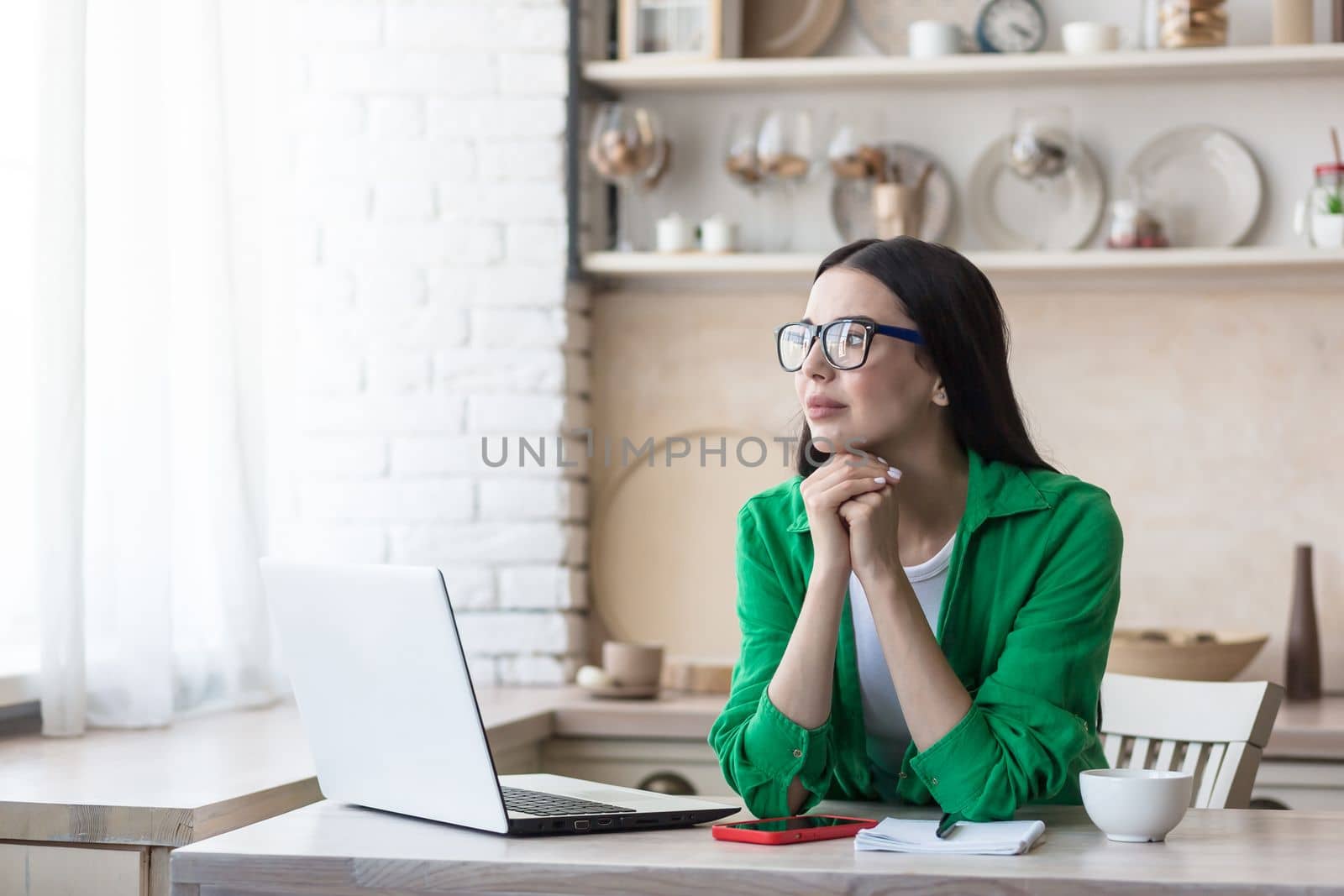 Upset young woman sitting at home in the kitchen in front of a laptop. Type on the keyboard, search, check. She looks thoughtfully out the window, resting her head on her hands.