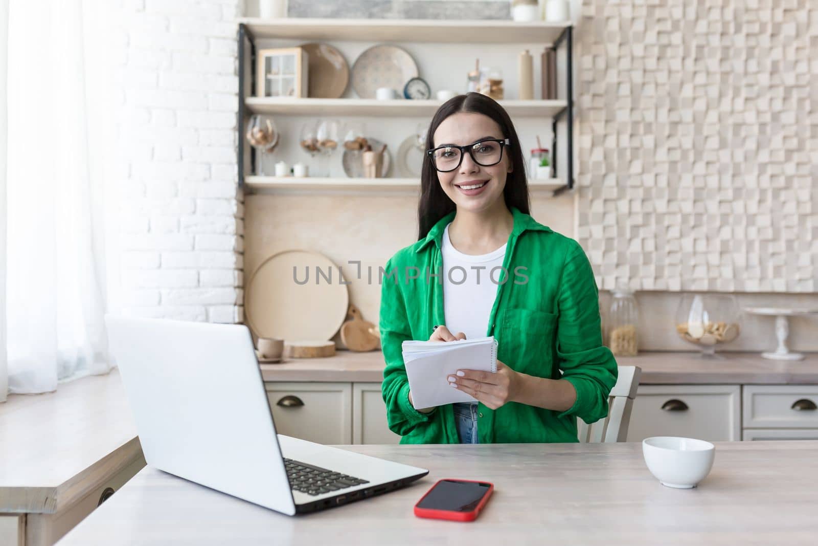 Portrait of a young woman teacher in glasses sitting at home in the kitchen with laptop. Works from home online. He is holding a notebook with a pen in his hands, smiling at the camera.