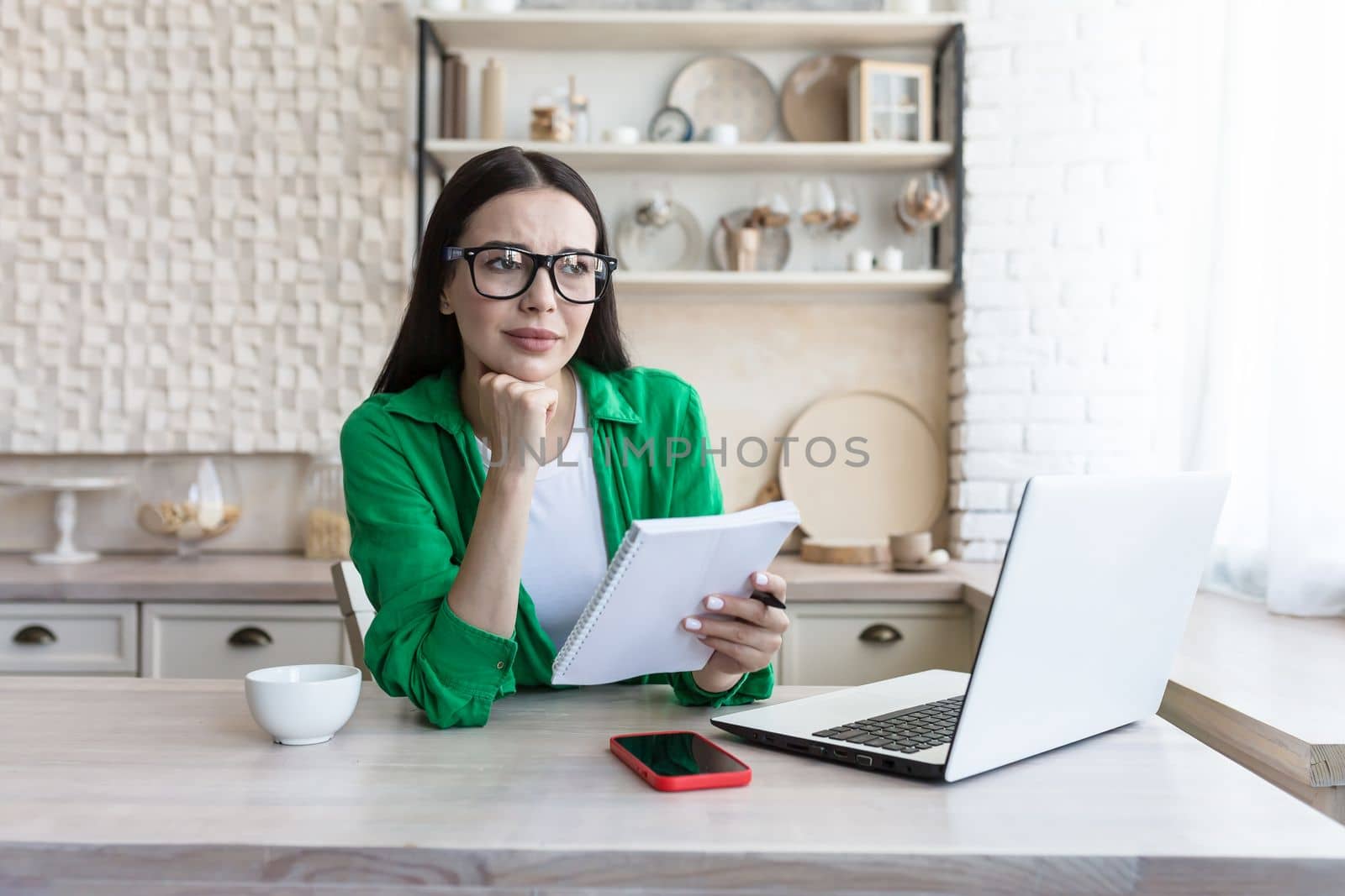 Upset young woman sitting at home in the kitchen in front of a laptop. Thoughtfully, she rested her head on her hand. Holds a notebook with a pen in his hand, checks documents, bills.