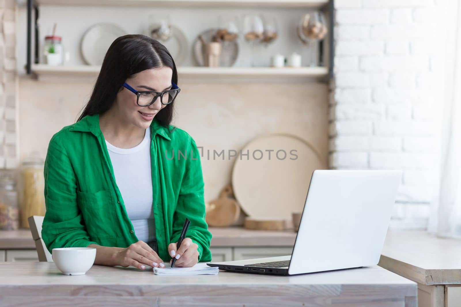 A young female student, a freelancer studies, works online, remotely at home from a laptop. He sits at the table, listens carefully to the lecture, writes with a pen in a notebook.