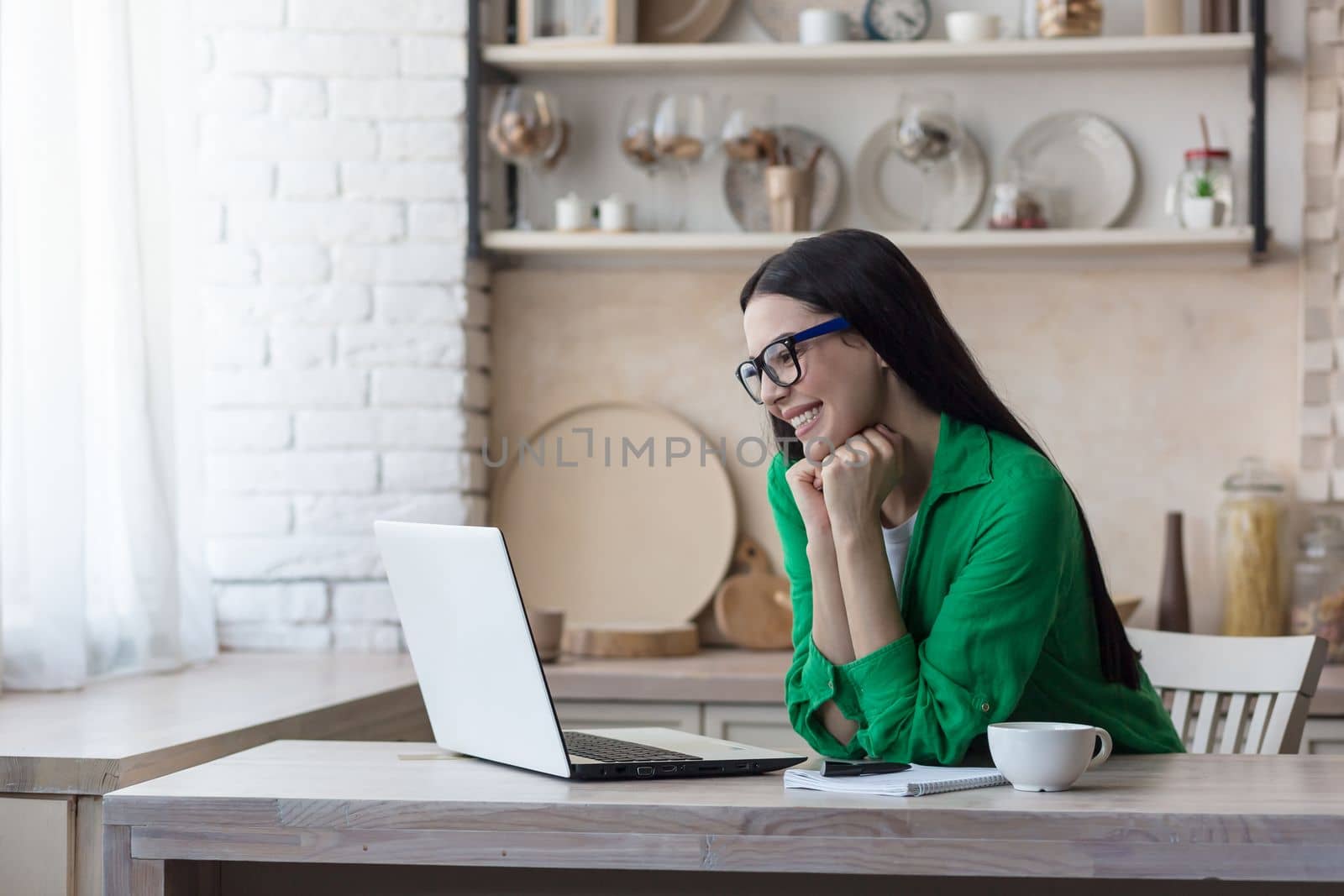 A happy young woman in quarantine sits at home in the kitchen and communicates with friends, parents, relatives through a video call from a laptop. Smiles, greets.